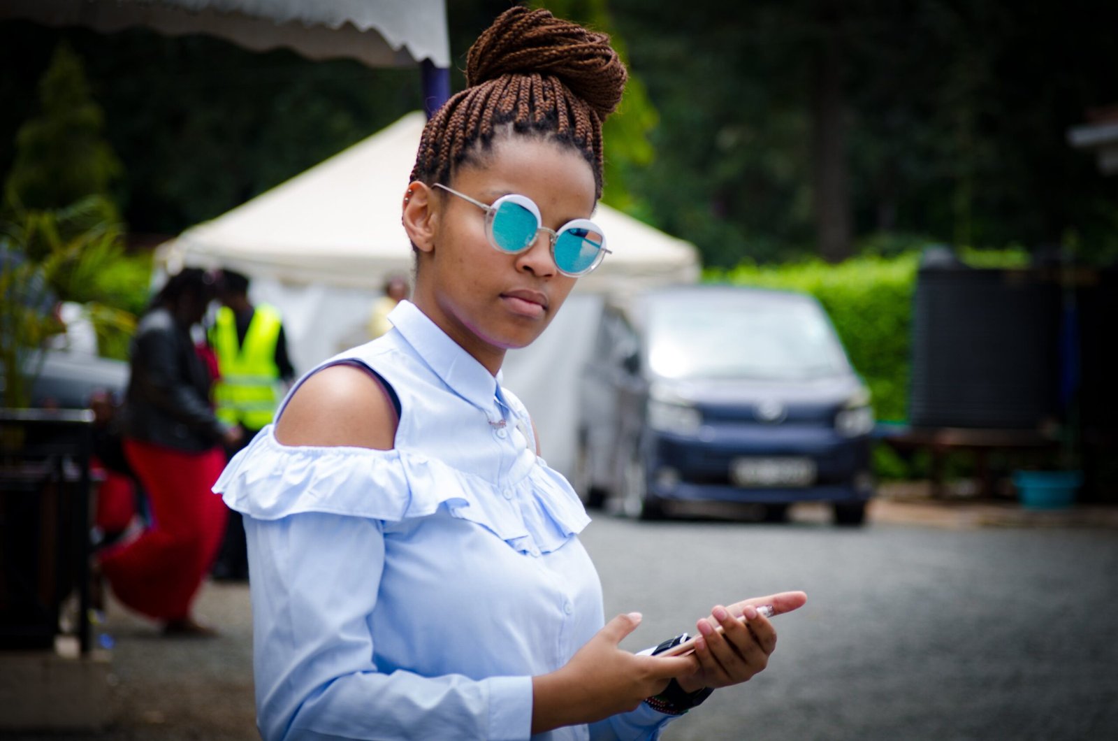 Stylish young woman wearing sunglasses and braided hair, holding a smartphone outdoors.